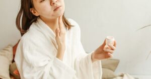 Wanita menyentuh dagunya mengenakan bathrobe putih saat rutinitas skincare pagi